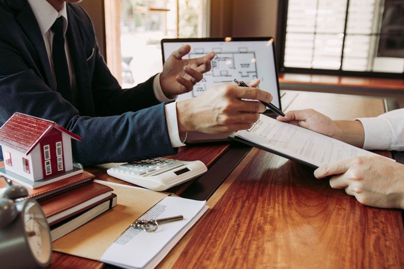 Two people discussing documents at a desk with a laptop, model house, calculator, and keys.